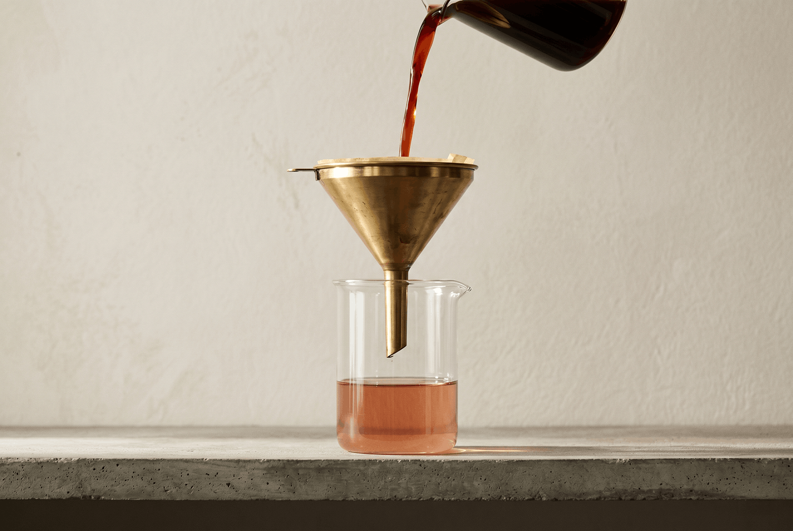 Dark liquid being poured through a brass funnel into a glass beaker, the filtered result lighter than the source